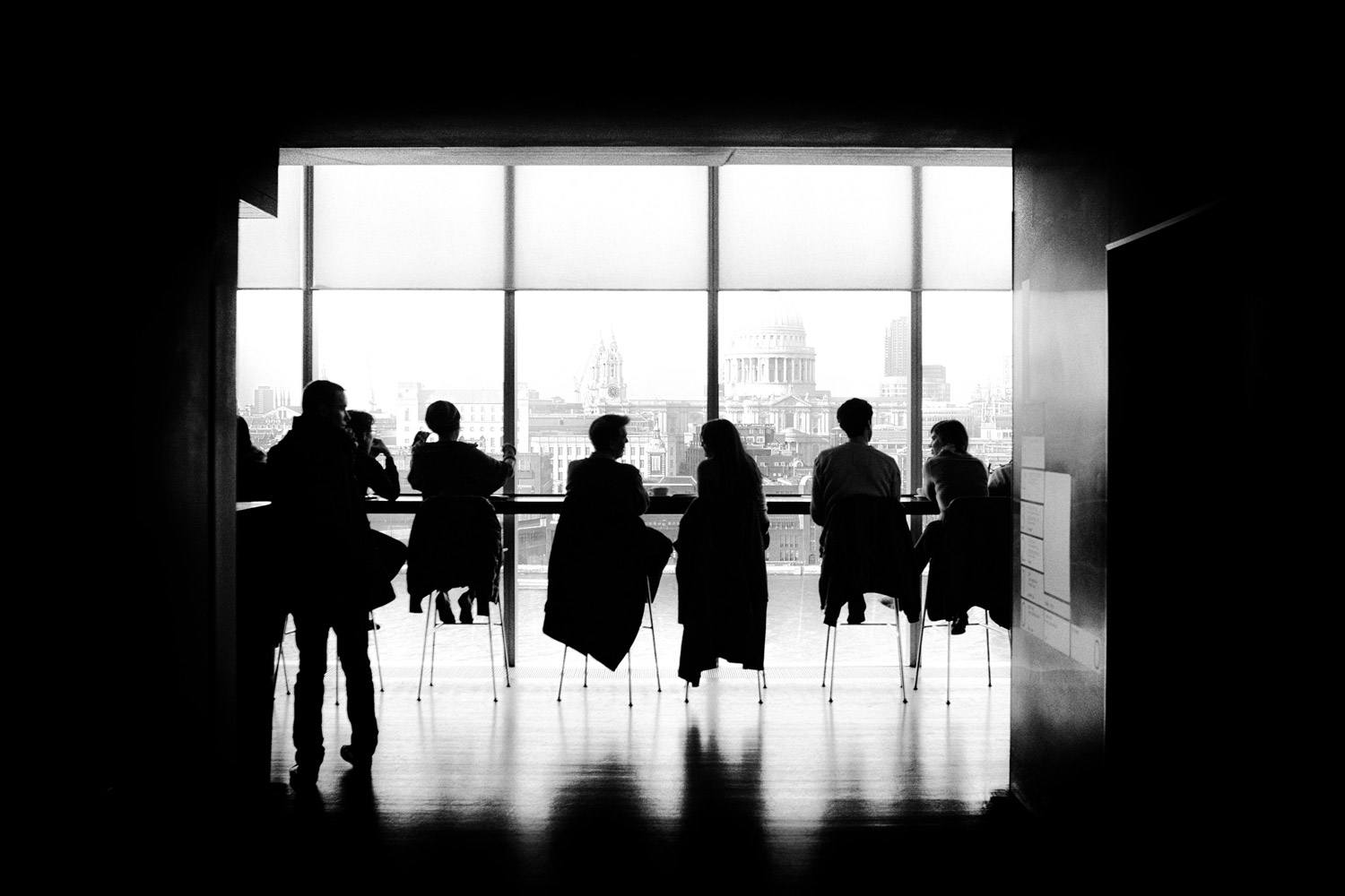 Black-and-white photo of 7 people engaged in conversation sitting in bar stools facing a window. Another man stands behind them.